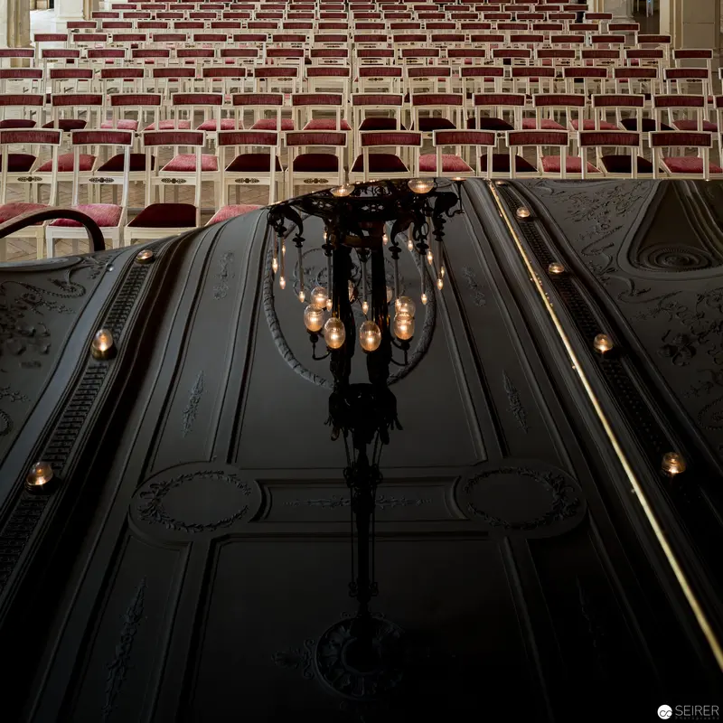 Ornate Lecture Hall Chandelier Illuminating Rows of Empty Burgundy Chairs