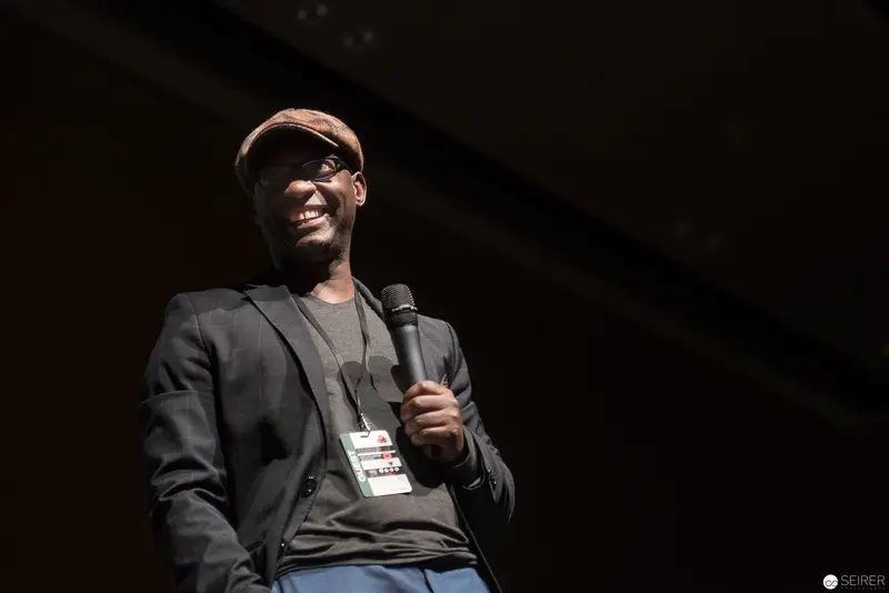 Smiling Speaker on Stage Wearing Flat Cap and Blazer