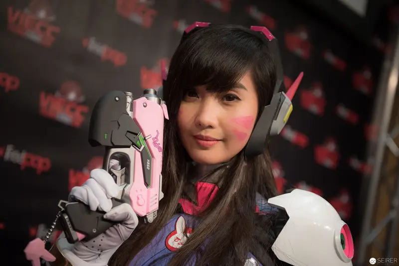 Cosplayer in D.Va Costume Posing with Prop Weapon at Convention Backdrop
