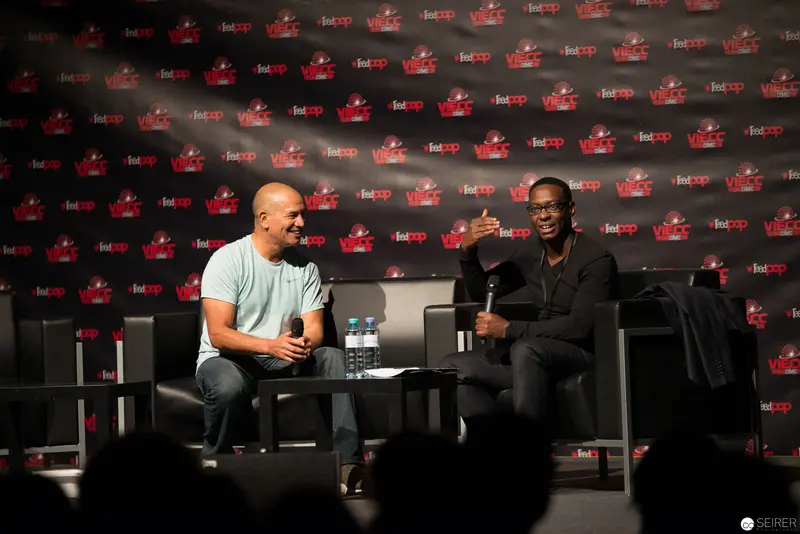 Two Speakers Engage in Panel Discussion at Public Event with Red Logo Backdrop