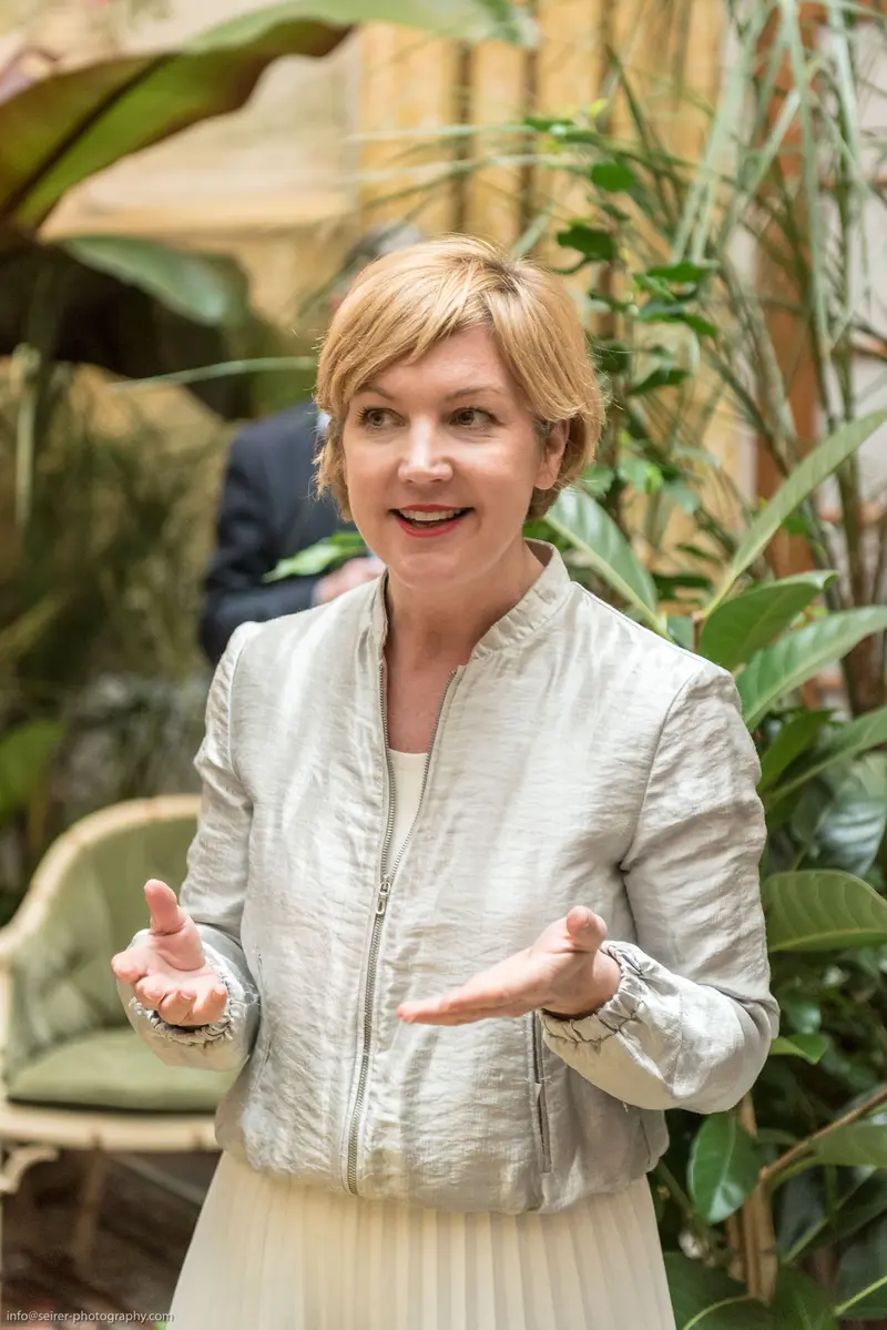 Woman in Silver Jacket Gesturing Amid Lush Indoor Greenery