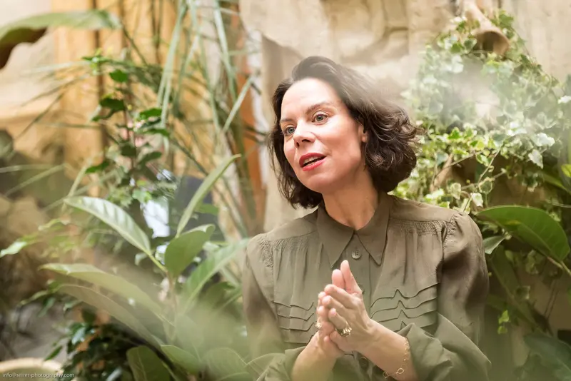 Woman in Olive Green Blouse Contemplating Among Lush Indoor Botanical Setting