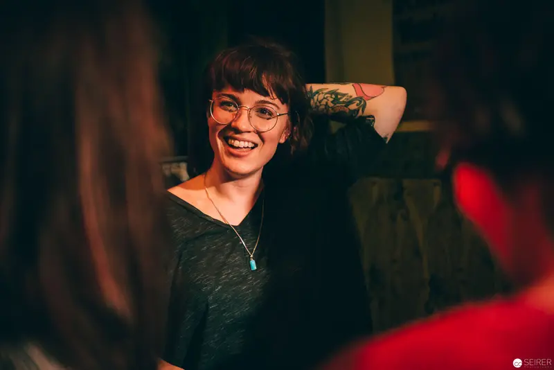 Smiling Person with Glasses and Tattoo in Warmly Lit Social Setting