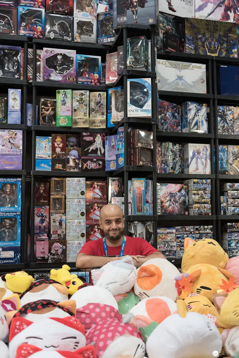 Manga Merchant Surrounded by Plush Characters in Densely Packed Collectibles Shop