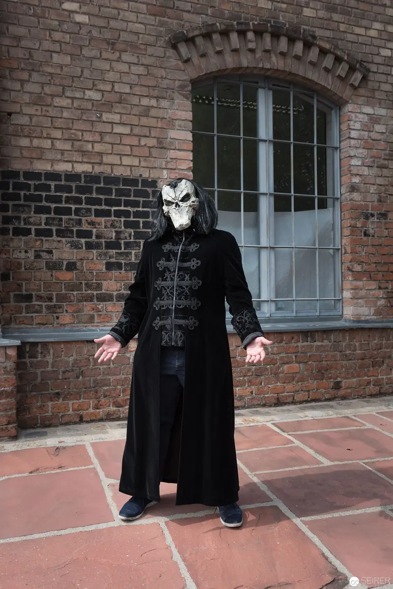 Masked Figure in Black Coat Standing Before Brick Wall and Arched Window