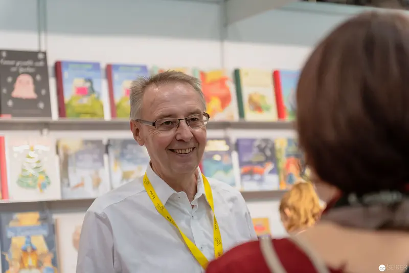Smiling Publisher Amidst Children's Book Display at Literary Event