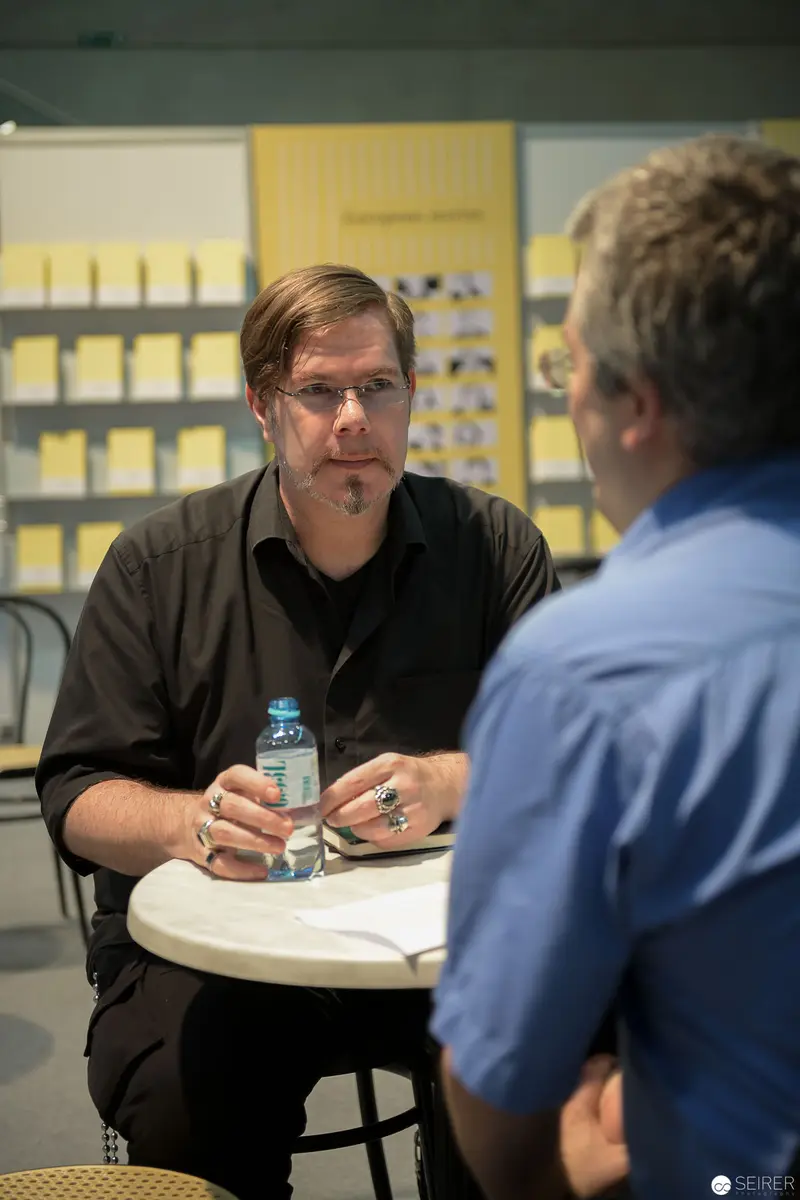 Two Men in Discussion at Conference Table with Post-It Notes Wall