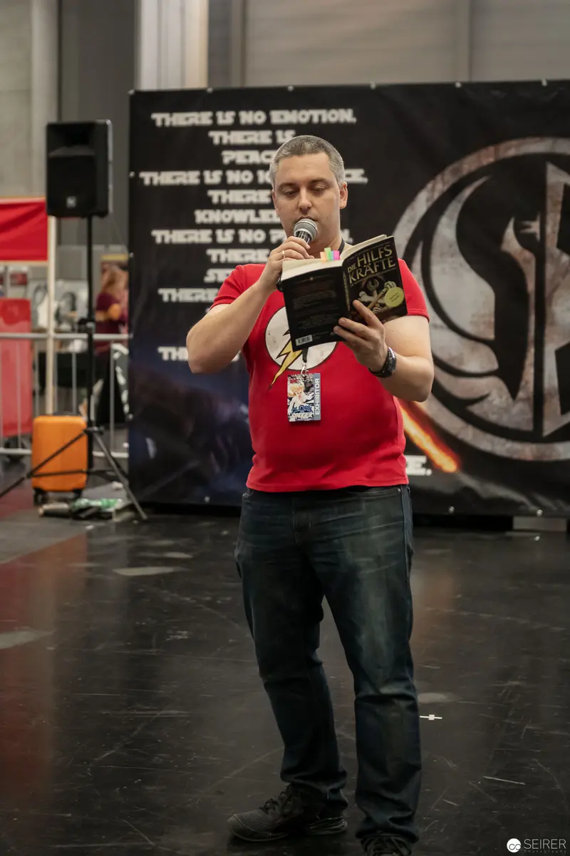 Speaker Presenting Book at Comic Convention Podium with Textual Backdrop
