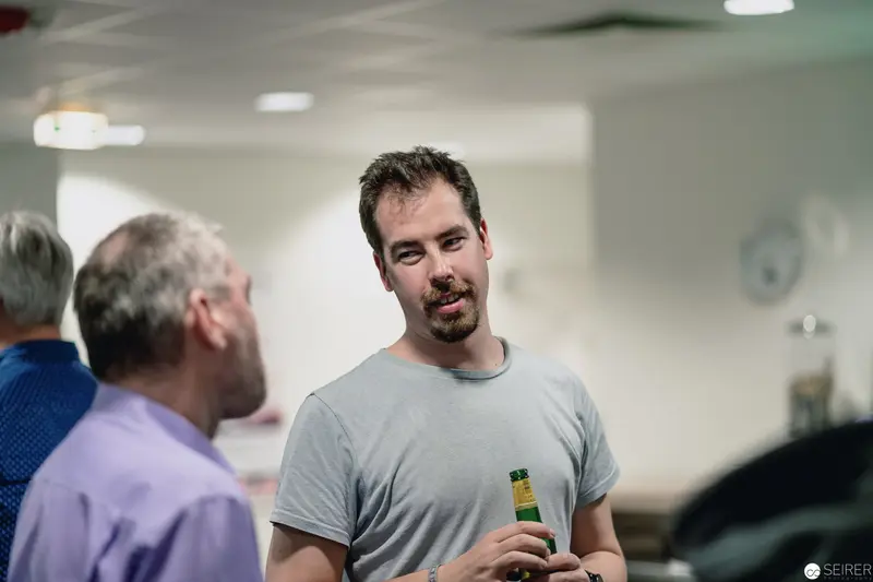 Beer-Holding Man in Gray Shirt Conversing with Colleagues in Indoor Setting