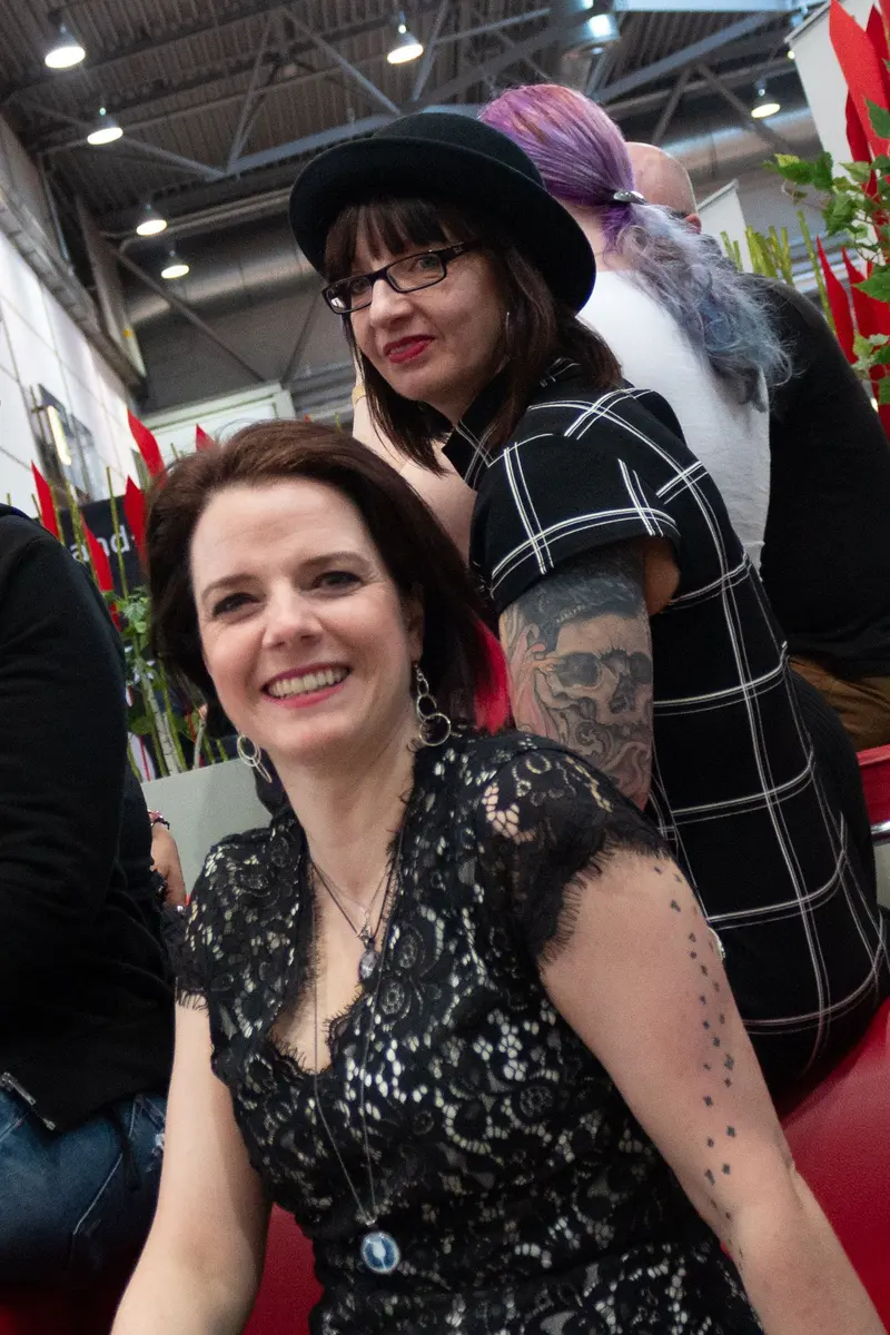 Two Women in Black at Indoor Event, Contrasting Styles and Expressions