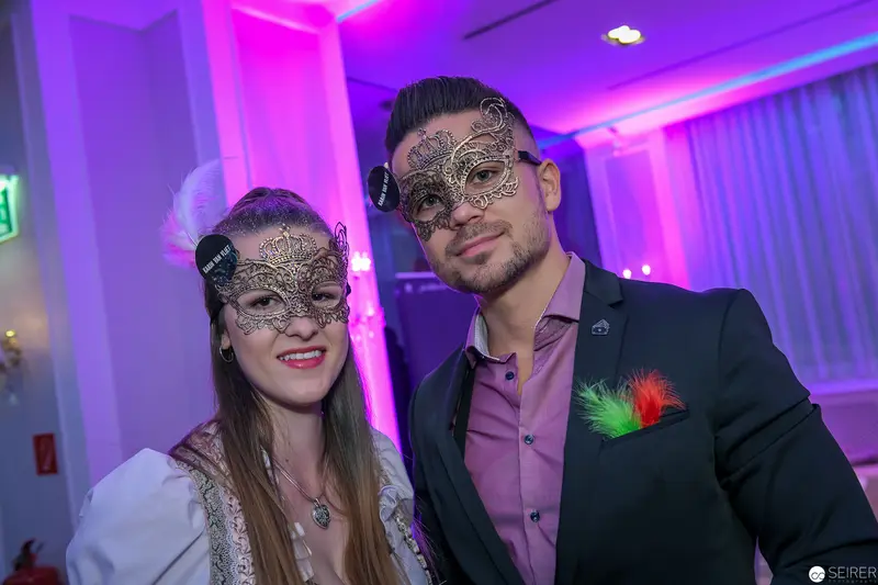 Masked Duo in Purple-Lit Event Space with Ornate Venetian-Style Masquerade Masks