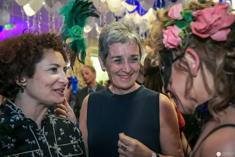 Women in Festive Attire Conversing at Social Gathering with Decorative Headwear