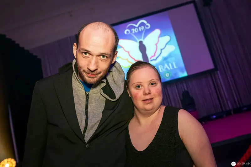 Two Individuals at Promotional Event with Butterfly Logo Backdrop