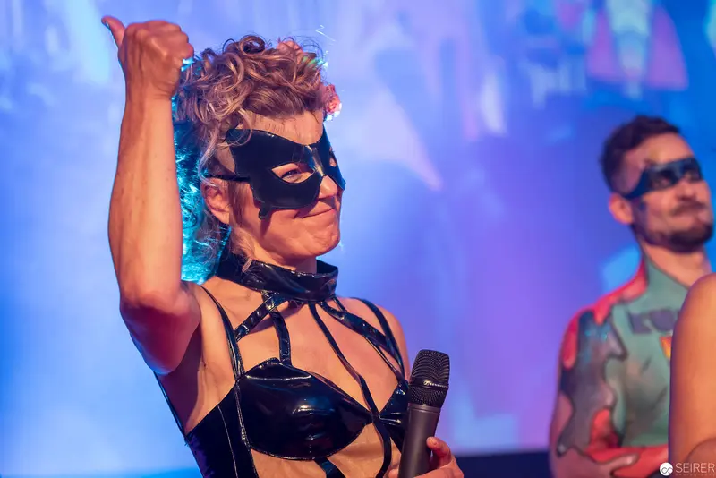 Performer in Black Latex Mask and Harness Gesturing on Stage with Microphone