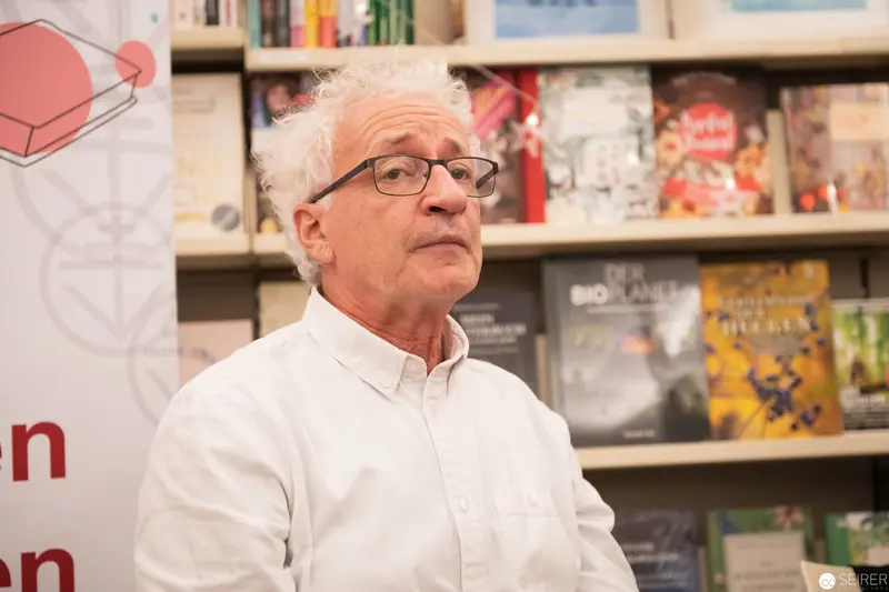 Older man in glasses speaking at book-lined venue event