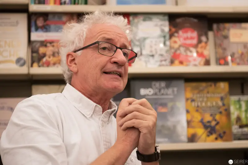 Older Man with White Hair Speaking at Bookshelf Event