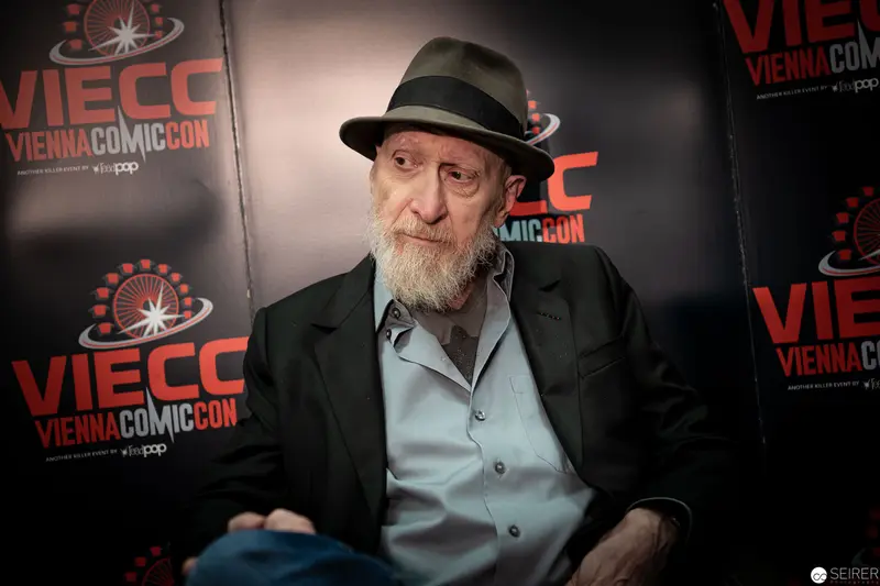 Seasoned Artist at Vienna ComicCon 2019, Seated Against Branded Backdrop