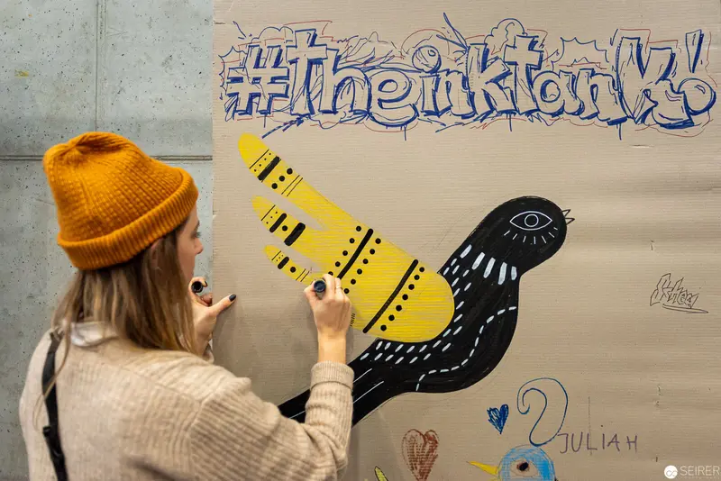 Artist Creating Stylized Bird Mural with #TheInkTank Hashtag in Background