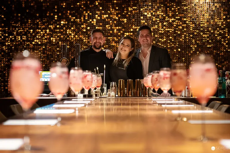 Bartenders Posing Behind Cocktail Bar with Sparkling Gold Sequin Backdrop