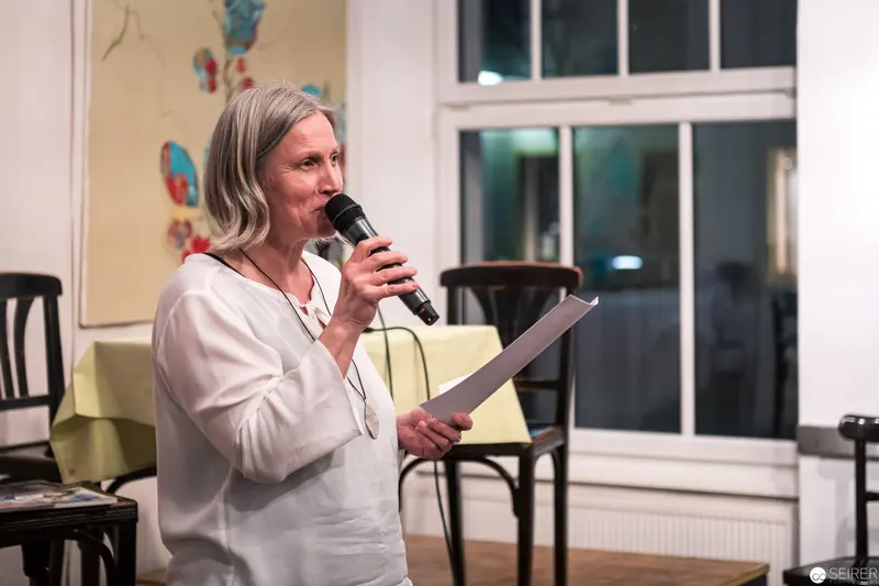 Woman Speaking at Indoor Event, Holding Microphone and Document