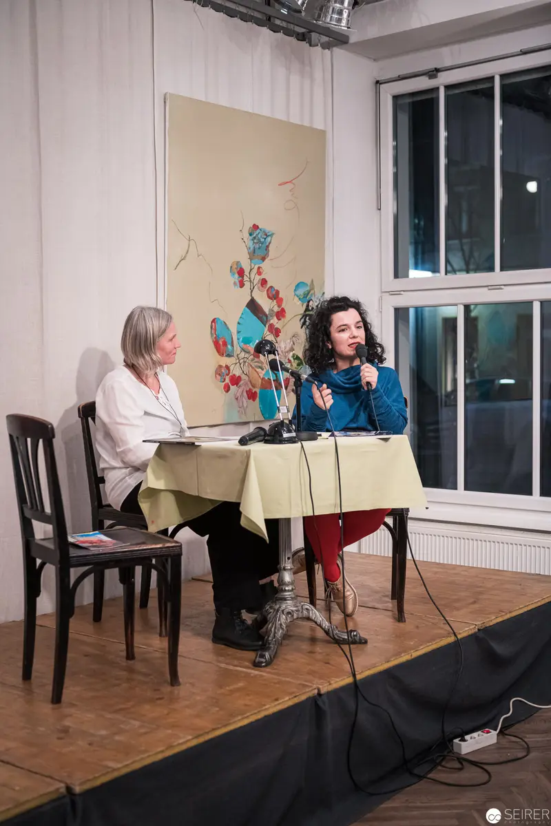 Two Women Engaged in Discussion at Public Panel Event with Artwork Background