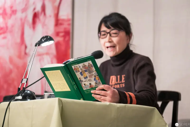Author Reading Aloud from Green Book Under Desk Lamp at Public Event