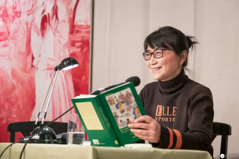 Author Reading at Public Event, Wearing Elle Sports Sweater and Glasses