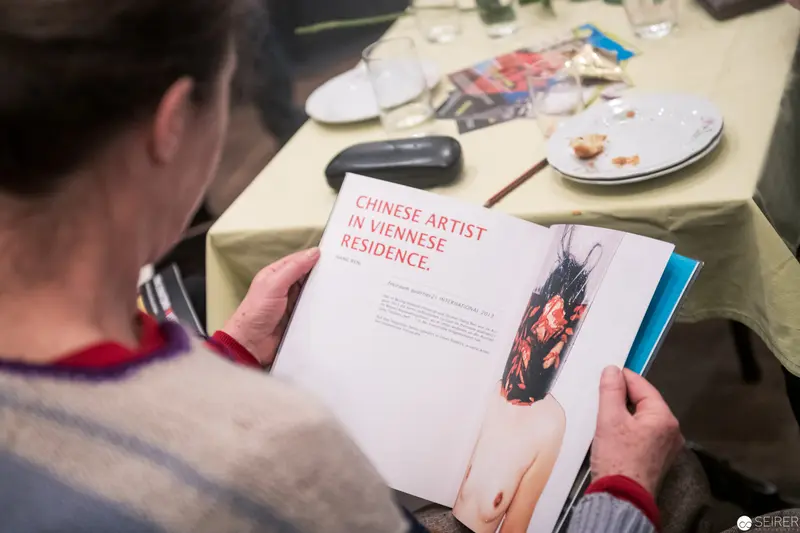Chinese Artist in Viennese Residence Examining Art Publication at Dining Table