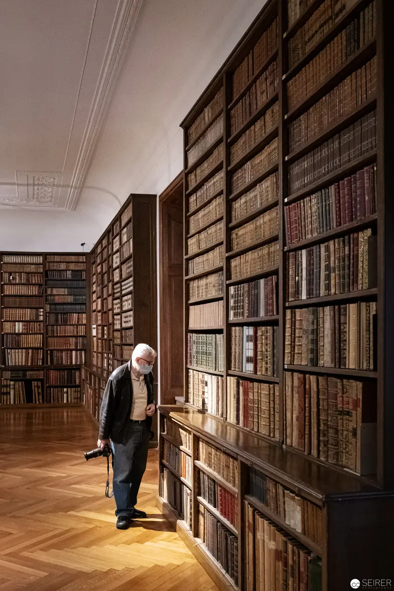 Elderly Scholar Exploring Vast Wooden Library Stacks with Walking Stick