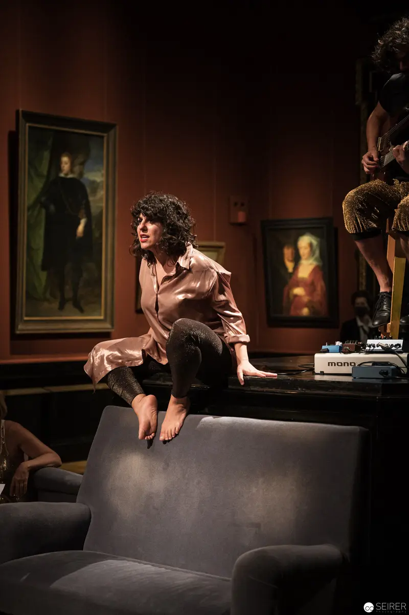 Performance Ganymed: Curly-Haired Performer Perched on Dark Furniture at KHM