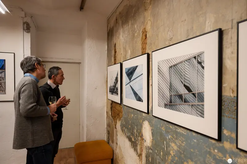 Two Viewers Examining Architectural Photographic Exhibition in Textured Gallery Space