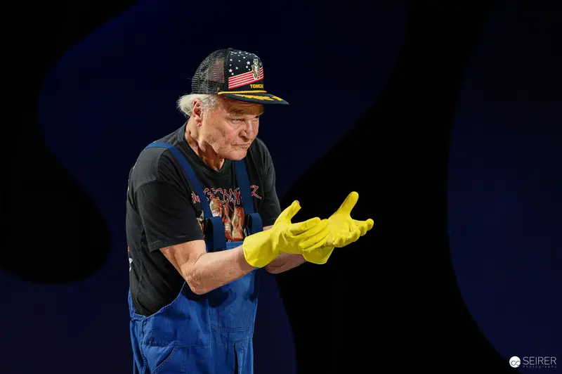 Frank Wedekind Wearing American Flag Cap and Yellow Rubber Gloves on Dark Stage