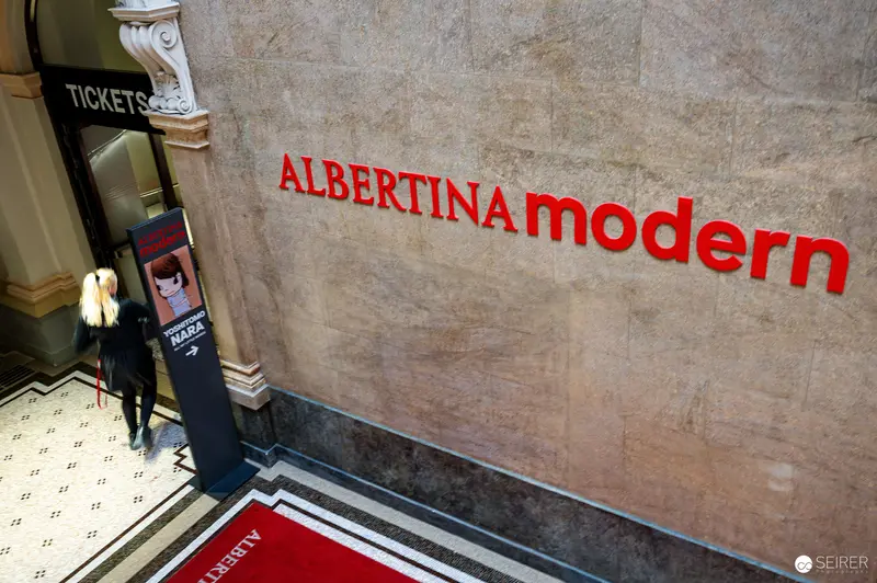 A visitor passes the ticket desk in the entrance foyer of Albertina Modern in Vienna, Austria, during the exhibition "Österreich – Deutschland. Malerei 1970–2020" in October 2023. Red "ALBERTINA modern" lettering is mounted on the stone wall beside a floor sign directing visitors to the concurrent Yoshitomo Nara exhibition.