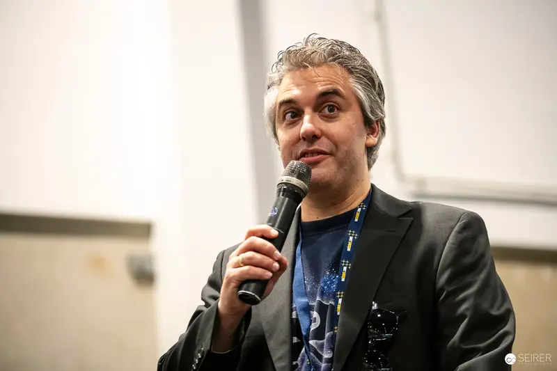 Comic Con Speaker in Black Blazer Addresses Audience with Dynamic Microphone Gesture