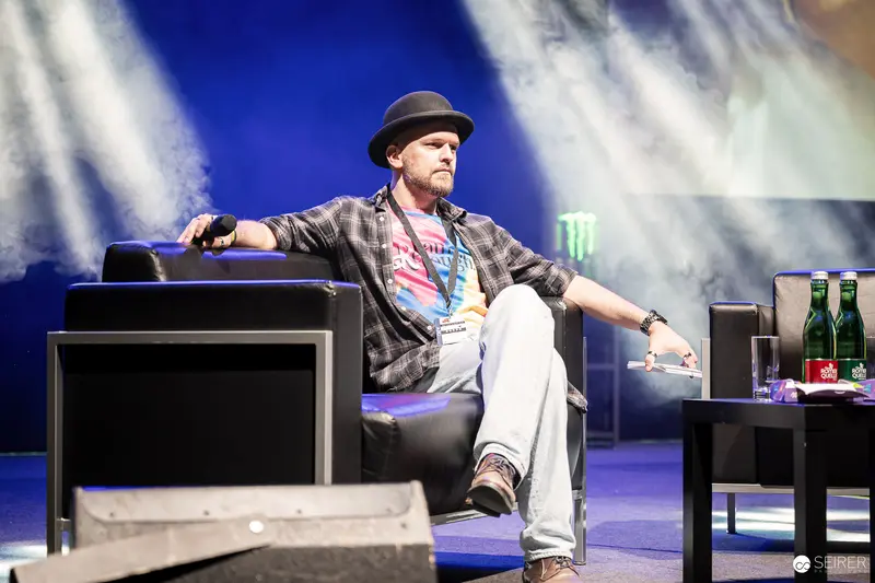 Comic Con Presenter in Plaid Jacket and Hat Seated on Stage