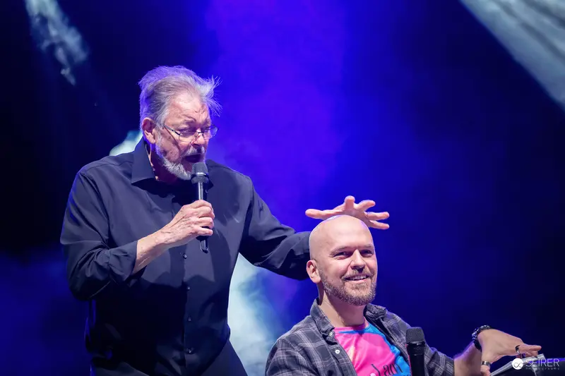 Panel guest with microphone gestures over seated moderator's head at VIECC Vienna Comic Con 2023