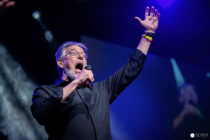 Grey-bearded panel host with raised arm shouts into microphone on VIECC Vienna Comic Con stage