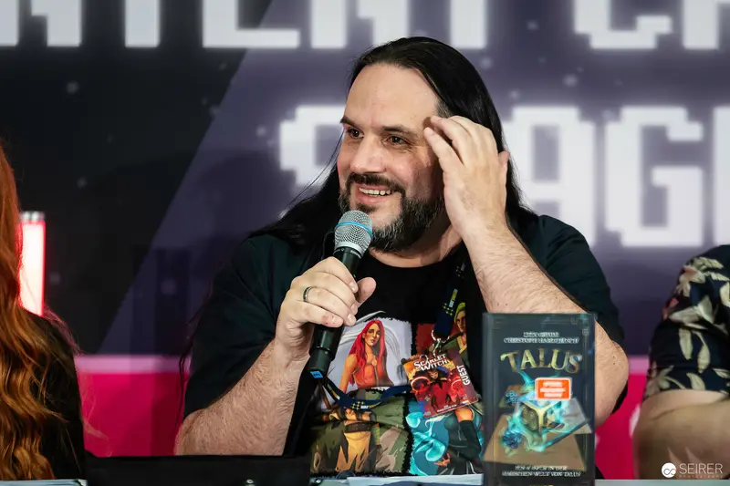 Long-haired panellist with microphone beside "Talus" novel at VIECC Vienna Comic Con 2023