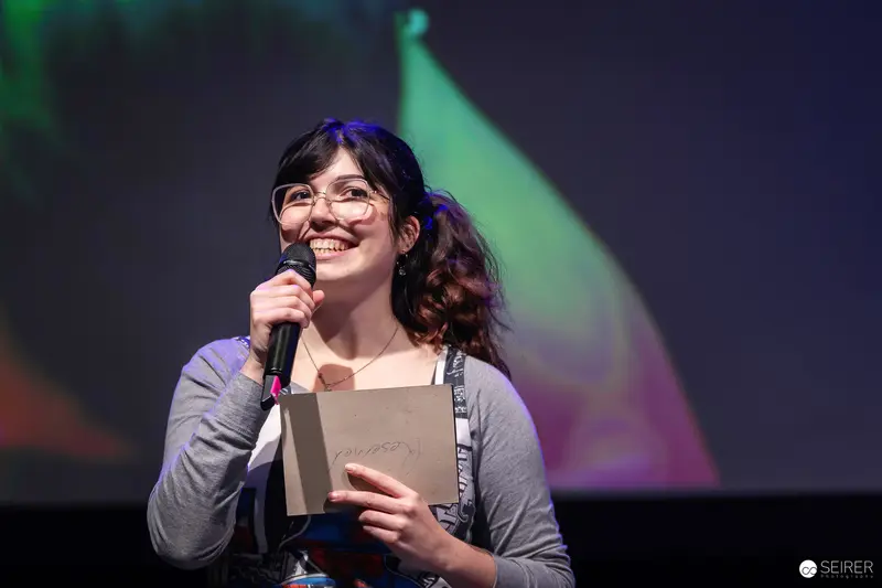 Smiling stage host with handheld microphone and cue card at VIECC Vienna Comic Con 2023