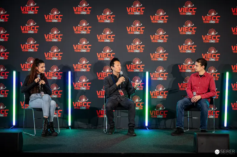 Three-person panel discussion on VIECC stage with Vienna Comic Con backdrop, November 2023