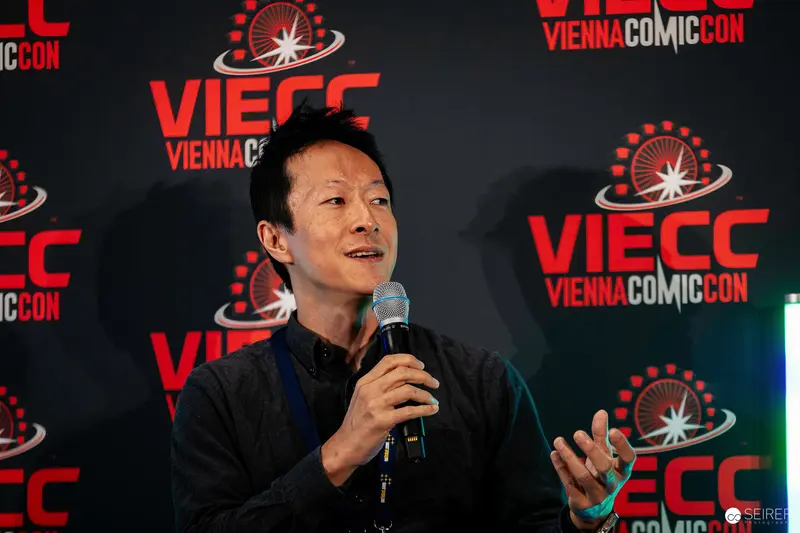Panel guest speaking into handheld microphone at VIECC Vienna Comic Con 2023 branding wall