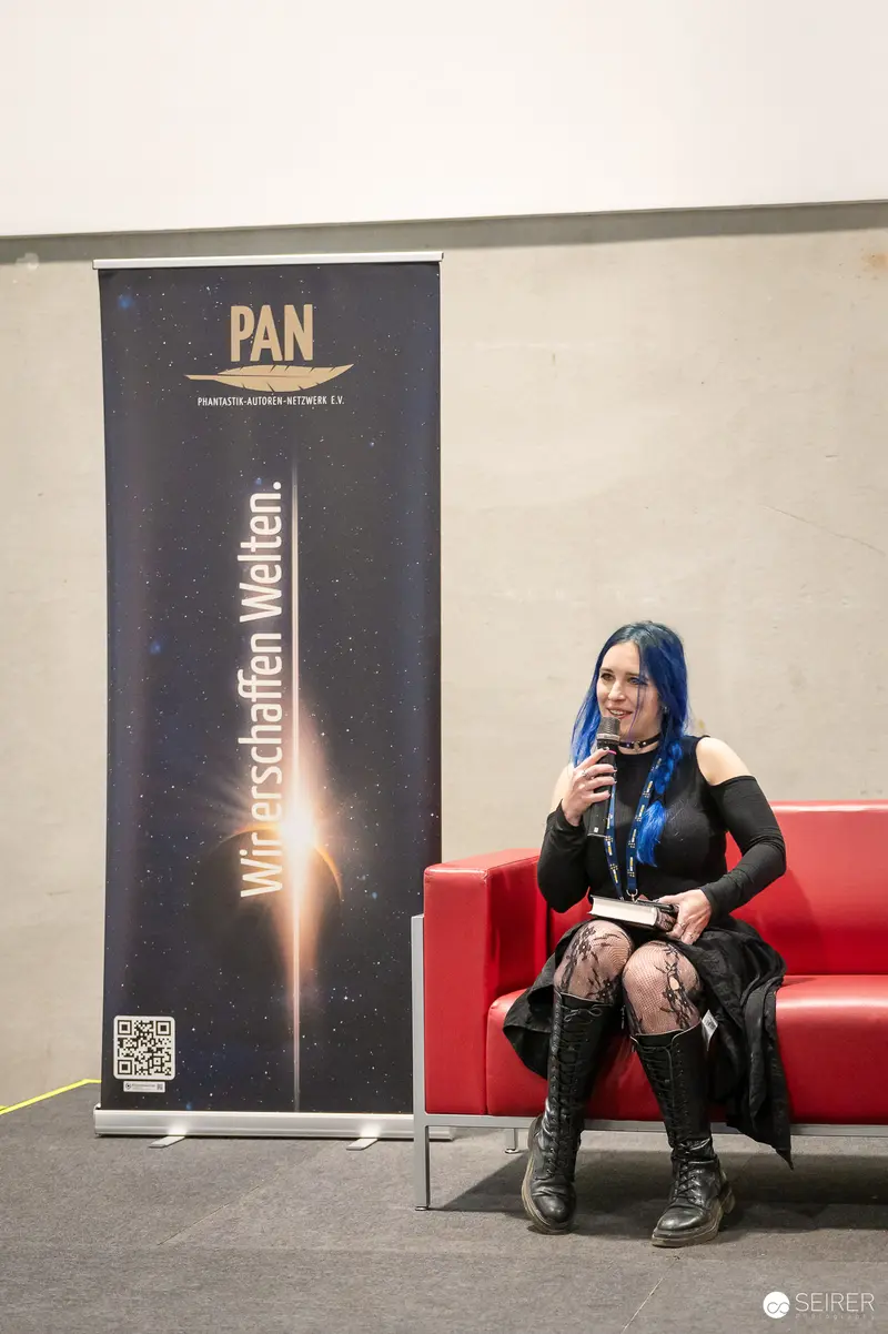 Blue-haired author speaking at PAN Phantastik-Autoren-Netzwerk panel, Vienna Comic Con 2023