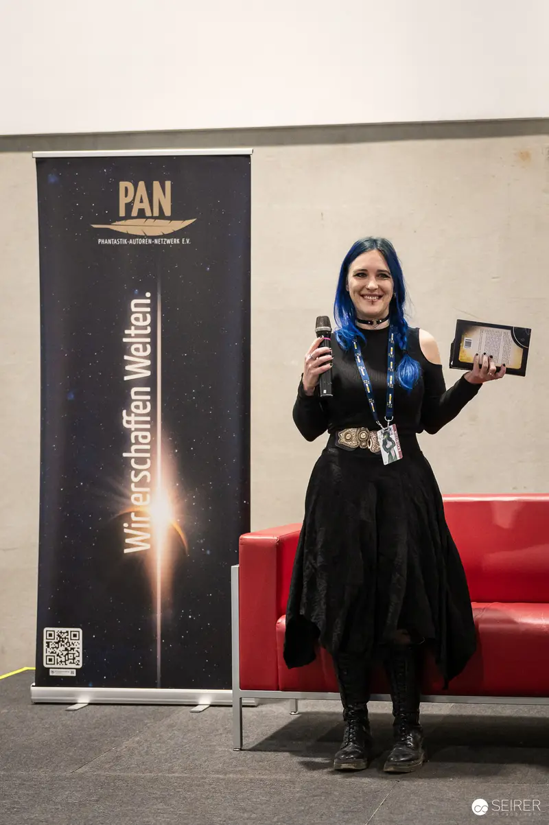 Blue-haired author presents book beside PAN Phantastik-Autoren-Netzwerk banner at Vienna Comic Con 2023
