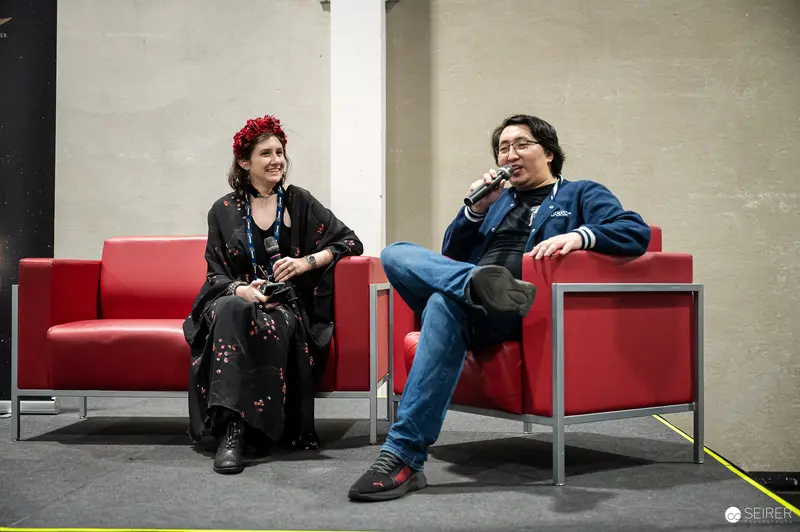 Panel guest speaking into microphone beside moderator on red armchairs at VIECC Vienna Comic Con 2023