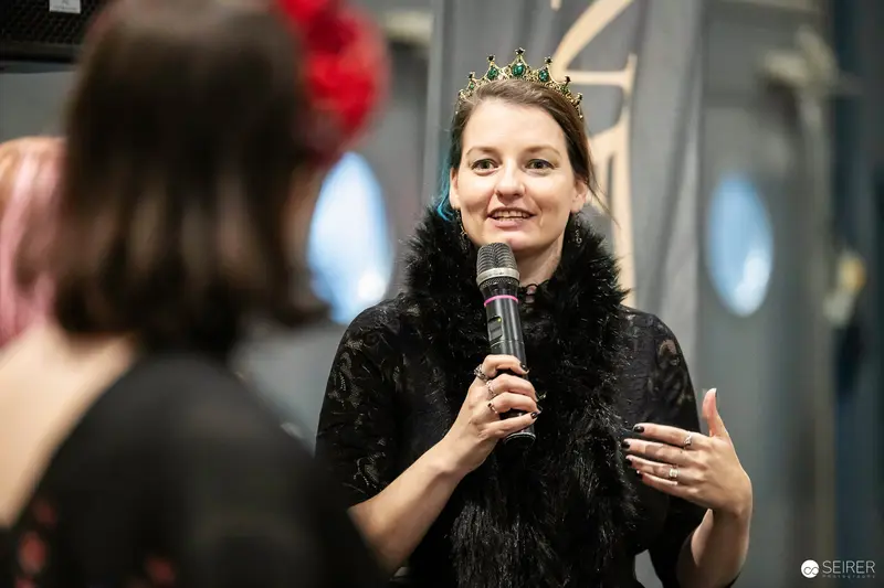 Cosplay host in emerald crown and black feather boa speaks on stage at VIECC 2023