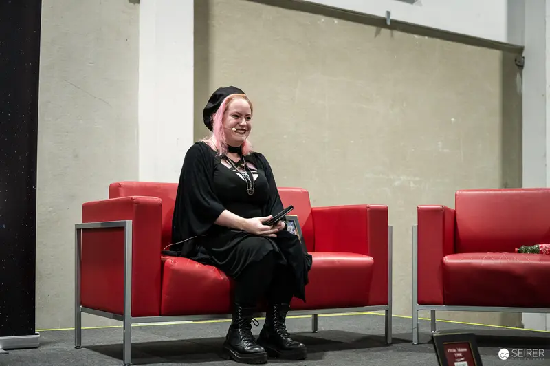 Panel speaker with pink hair and black beret seated on red armchair, VIECC Vienna Comic Con 2023