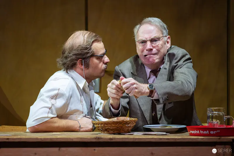 Der Himbeerpflücker Theater Scene: Two Men at Wooden Table in Intense Interaction