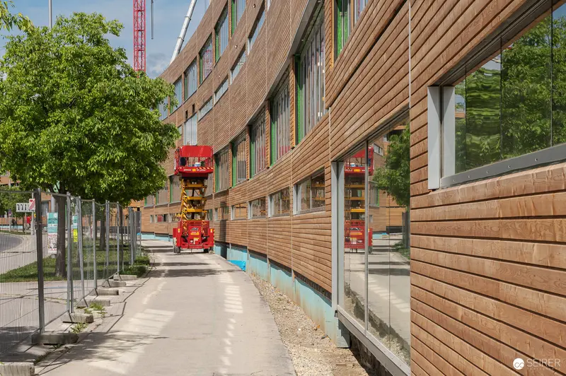 Modern Wood-Clad Building Under Construction with Red Cranes and Visitor Walkway