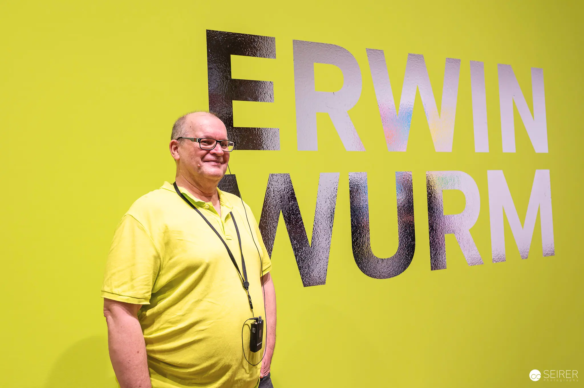 Erwin Wurm Stands Beside Large Text Display at Personal Exhibition