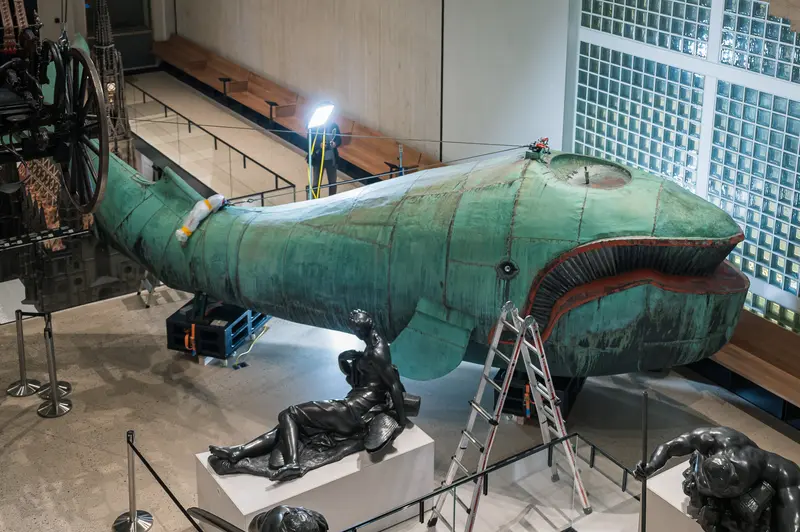 Maria Benke's 10-meter copper whale sculpture "Poldi" undergoes conservation work during the Wien Museum's 2024 Tage der Restaurierung program, its distinctive green patinated surface and red-painted lips visible as industrial maintenance equipment and ladders support the restoration process. The sculpture, originally salvaged from the demolished Gasthaus zum Walfisch, rests on support structures while museum visitors observe the behind-the-scenes work from upper-level walkways. The wooden frame and copper sheeting construction demonstrates traditional Viennese craftsmanship in this cultural artifact's preservation.